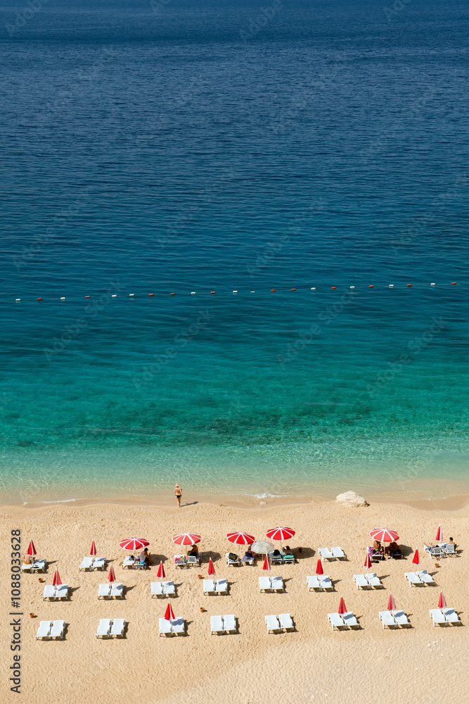 An aerial view of a beautiful beach with turquoise waters, featuring rows of sun loungers and red umbrellas neatly arranged on the sandy shore. An aerial view of a beautiful beach with turquoise waters, featuring rows of sun loungers and red umbrellas neatly arranged on the sandy shore.
