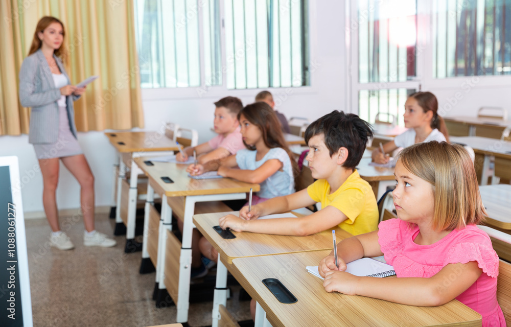 Schoolkids sitting at desks in classroom. They're listening to feamle teacher who's explaining them lesson information. Schoolkids sitting at desks in classroom. They're listening to feamle teacher who's explaining them lesson information.