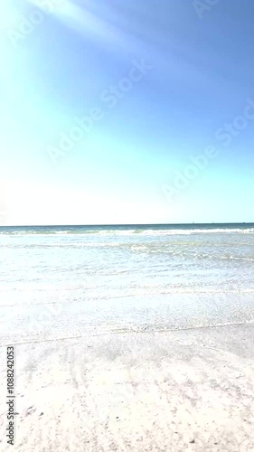 Serene ocean view with gentle waves lapping at a sandy shore under a clear blue sky. Bright sunlight illuminates the water, creating a tranquil and refreshing beach atmosphere.