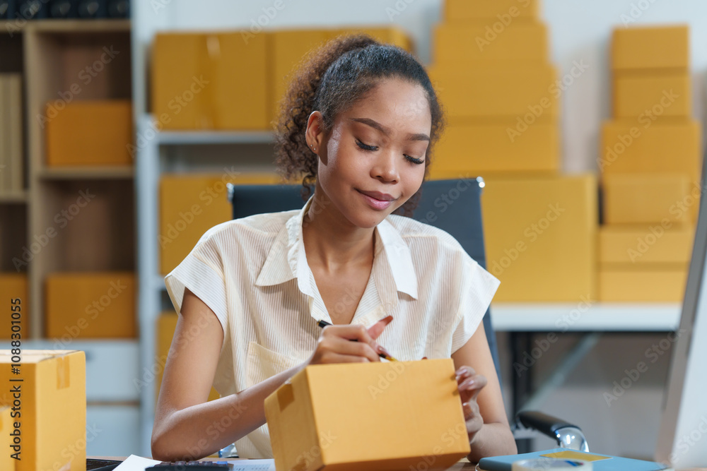 A beautiful African American woman sits at a desk, checking items, packing products, taking orders, monitoring sales on her computer. Packages are ready for shipping, showcasing her thriving online A beautiful African American woman sits at a desk, checking items, packing products, taking orders, monitoring sales on her computer. Packages are ready for shipping, showcasing her thriving online