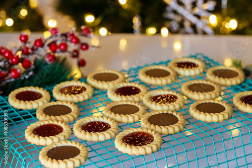 Cookies with chocolate and jam fillings cool on a wire rack surrounded by holiday decorations and soft lights. The warm atmosphere invites festive cheer during the season.