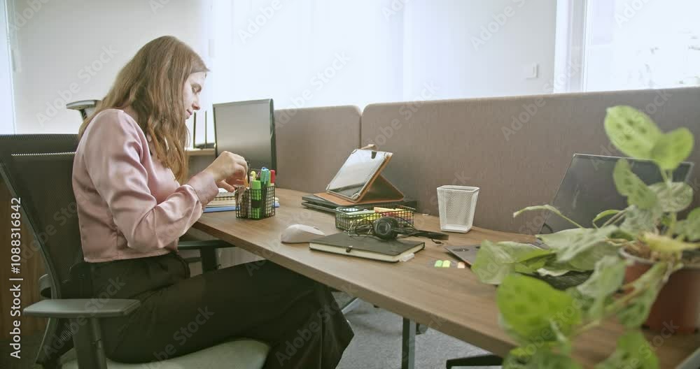 A young woman sits at her desk in a modern office, organizing her colorful stationery. She takes notes in her notebook. Natural light illuminates the workspace, creating a productive atmosphere. A young woman sits at her desk in a modern office, organizing her colorful stationery. She takes notes in her notebook. Natural light illuminates the workspace, creating a productive atmosphere.