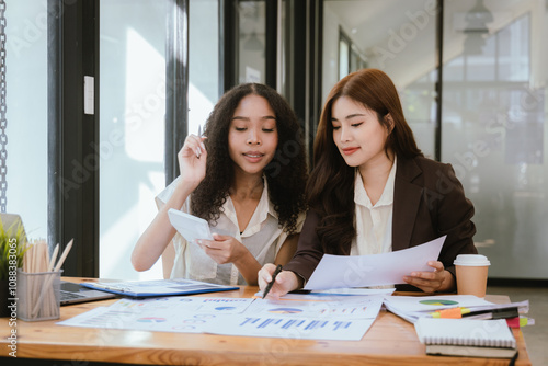 Two Asian businesswomen in suits talking at work, discussing chart analysis, business investment.