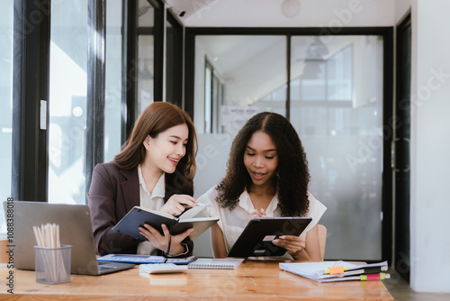 Two Asian businesswomen in suits talking in a modern workplace. Thai women, Southeast Asians, looking at laptops together.