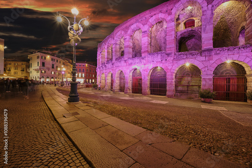 The Arena di Verona, illuminated in fuchsia hues against a vibrant pink sunset sky. Long exposure evening shot capturing the motion blur of people passing by.