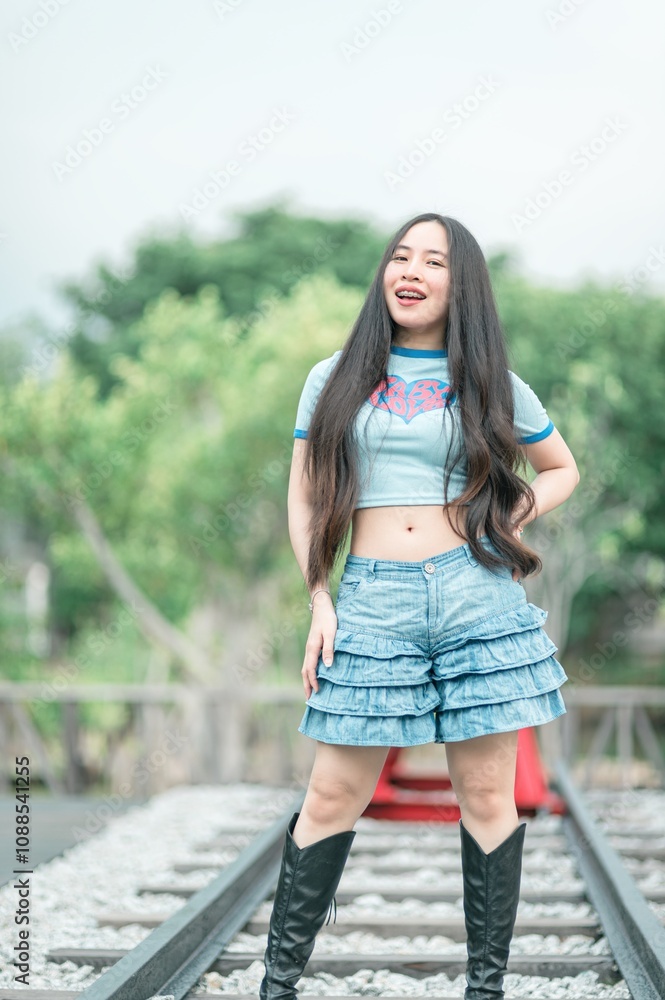 Stylish Young Woman Posing on Railway Tracks in Trendy Outfit, Featuring Denim Shorts and Knee-High Boots, Surrounded by Lush Greenery Stylish Young Woman Posing on Railway Tracks in Trendy Outfit, Featuring Denim Shorts and Knee-High Boots, Surrounded by Lush Greenery