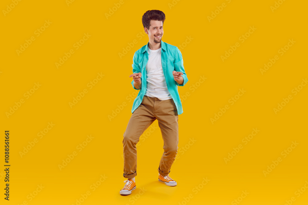 Hey you, yes you. You're going to the party. Full body length happy handsome young man in casual turquoise shirt and brown pants standing on yellow background, pointing fingers at you and smiling Hey you, yes you. You're going to the party. Full body length happy handsome young man in casual turquoise shirt and brown pants standing on yellow background, pointing fingers at you and smiling
