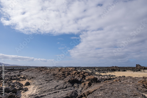 White sand beach with solidified volcanic lava flows, black rocks. Mountains in the background and sky with big white clouds. Caletón Blanco, Lanzarote, Canary Islands, Spain