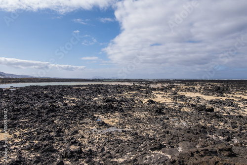 White sand beach with solidified volcanic lava flows, black rocks. Mountains in the background and sky with big white clouds. Caletón Blanco, Lanzarote, Canary Islands, Spain