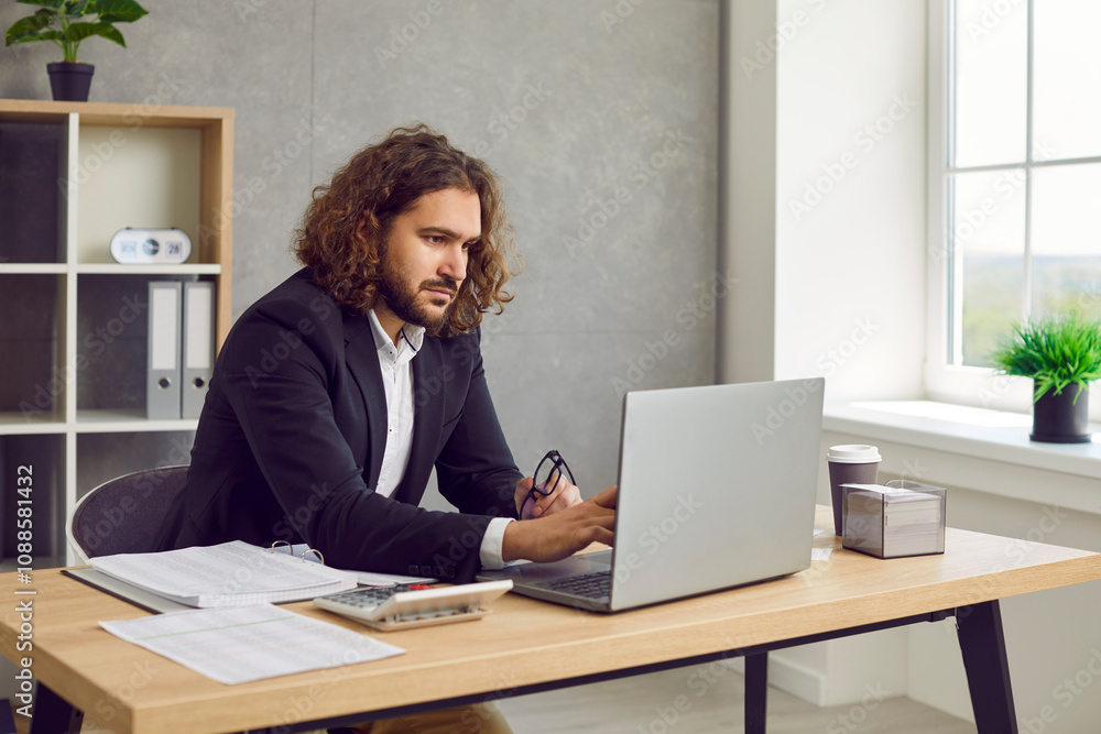 Portrait of confident young attractive business man with long hair in suit looking seriously at laptop computer monitor screen working at the desk on his workplace and typing in the office. Portrait of confident young attractive business man with long hair in suit looking seriously at laptop computer monitor screen working at the desk on his workplace and typing in the office.