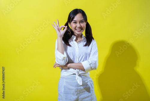 Asian woman smiling brightly, showing an "OK" hand gesture with confidence. She is dressed in a white shirt and striped pants, standing against a vibrant yellow background.