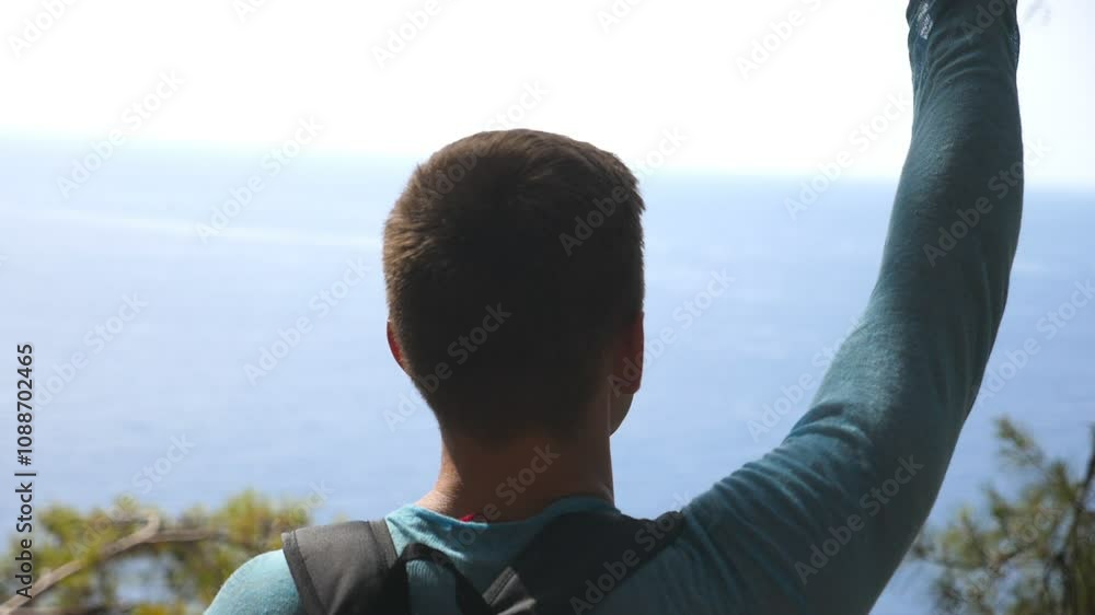 Cheerful hiker with backpack rejoicing success on edge of mountain victoriously raising hand. Male tourist actively resting during trip and enjoying freedom. Beautiful landscape with sea view. Slow mo Cheerful hiker with backpack rejoicing success on edge of mountain victoriously raising hand. Male tourist actively resting during trip and enjoying freedom. Beautiful landscape with sea view. Slow mo