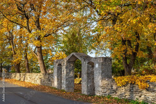 A rustic stone wall of Joelahtme church (Estonian - Jõelähtme kirik) stretches alongside a road, with a traditional stone archway. Above, large trees with bright golden-yellow leaves frame the scene. 