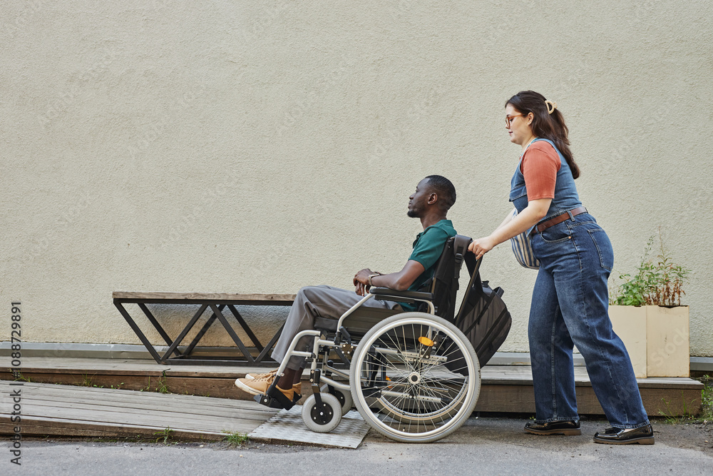 Obraz premium Minimal full length shot of young African American man with disability moving up on ramp with friend or partner assisting copy space