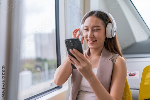 Asian Woman Using Phone and Headphones Listening To Music and Playing Games While Commuting To Work in a Public Train