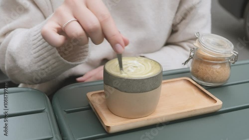 Wallpaper Mural A person in a warm sweater stirs a frothy matcha latte, creating a calming ambiance. The ceramic mug sits on a wooden tray beside a jar of sugar, on a green table Torontodigital.ca