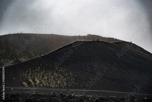 A dormant crater of Mount Etna viewed from the distance, with tiny silhouettes of people walking along its edge. Stock photo photo