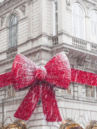 A huge red bow with a ribbon on the facade of a building in Christmas lights during a snowfall.