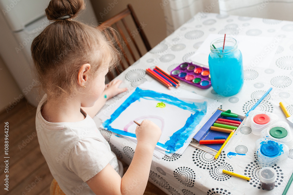 A cute girl draws with paints and pencils at a table near the window. A cute girl draws with paints and pencils at a table near the window.