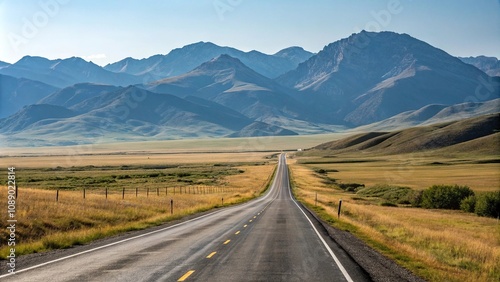 A scenic drive on a straight asphalt road through a vast open landscape with rolling hills and towering mountains visible beyond the horizon, isolation, wide open spaces, solitude, serenity, blue sky