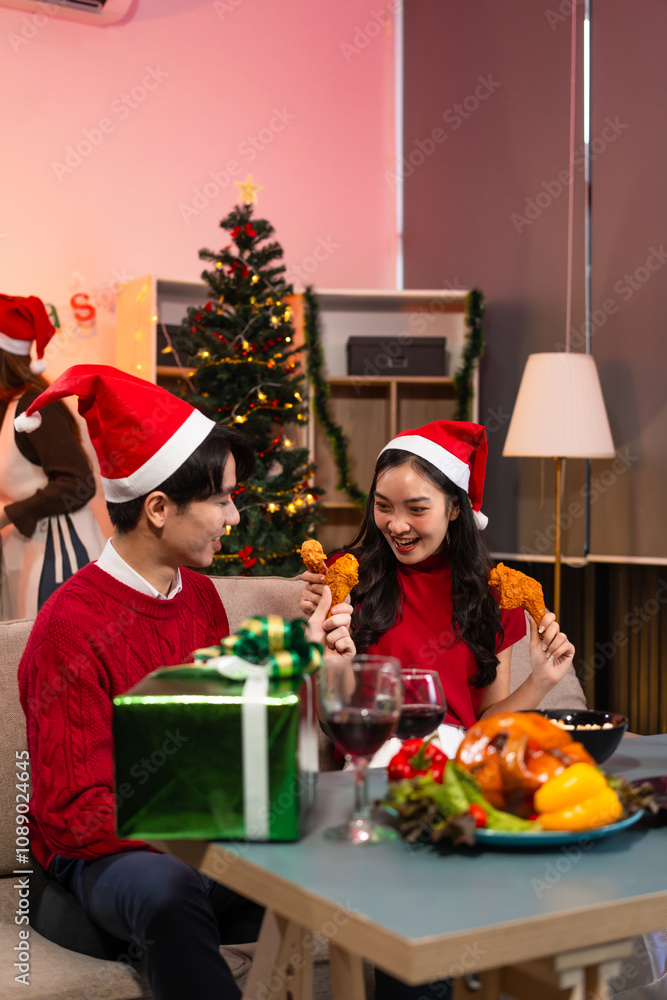 Two handsome young men and two beautiful young women wearing Christmas hats laughing joyfully at a Christmas party with delicious food, drinks and a cheerful exchange of thoughtful gifts
