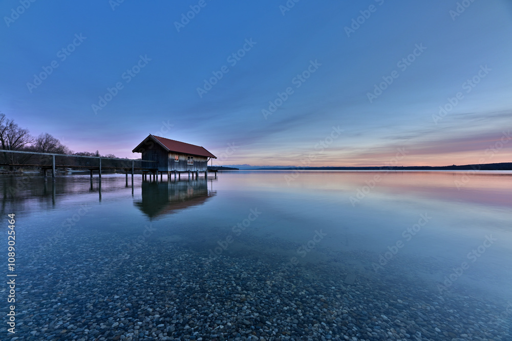 Ein Bootshaus steht einsam im absolut stillen und klaren Wasser eines See. Ein schönes Abendrot zeichnet sich am Himmel ab, und im klaren Wasser sieht man die Kiesel am Seegrund. Ein Bootshaus steht einsam im absolut stillen und klaren Wasser eines See. Ein schönes Abendrot zeichnet sich am Himmel ab, und im klaren Wasser sieht man die Kiesel am Seegrund.
