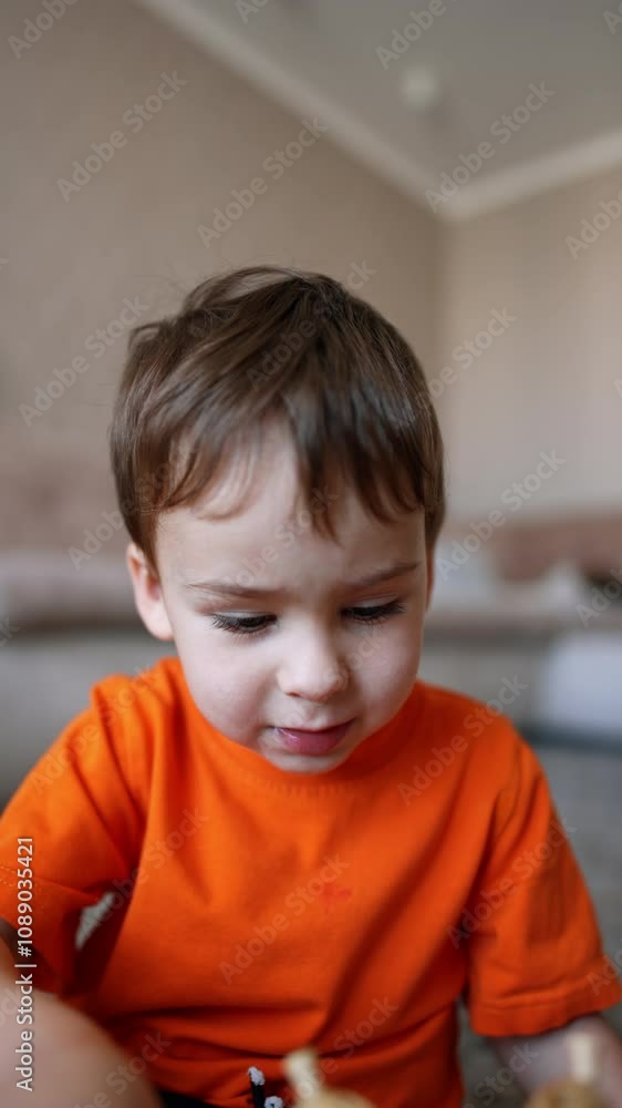 Beautiful Caucasian baby boy in orange t-shirt sits on the floor playing. Close portrait of a kid focused on a toy. Vertical video. Beautiful Caucasian baby boy in orange t-shirt sits on the floor playing. Close portrait of a kid focused on a toy. Vertical video.