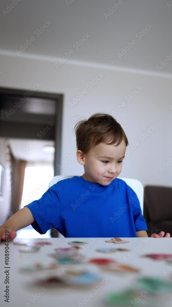 Happy smiling kid in blue t-shirt runs up to a desk. Toddler boy sits down at the desk and looks at the puzzles. Low angle view. Vertical video. Happy smiling kid in blue t-shirt runs up to a desk. Toddler boy sits down at the desk and looks at the puzzles. Low angle view. Vertical video.