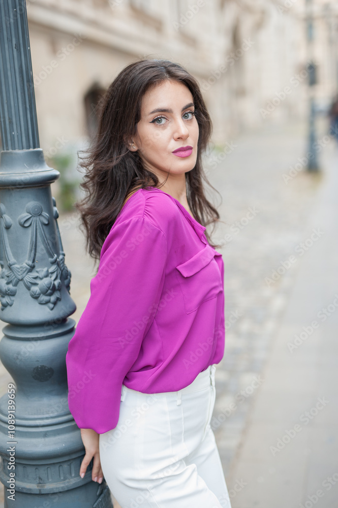 Beautiful young brunette woman in a purple blouse and white pants posing outdoor leans against a pillar .Portrait of a beautiful young brunette woman in a purple jacket posing