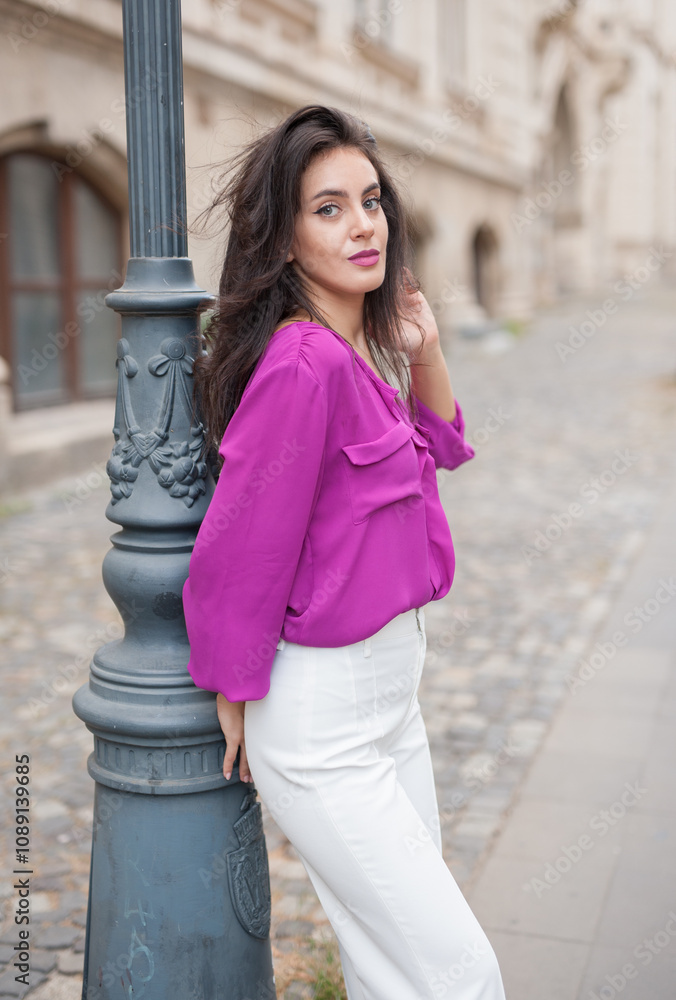 Beautiful young brunette woman in a purple blouse and white pants posing outdoor leans against a pillar .Portrait of a beautiful young brunette woman in a purple jacket posing