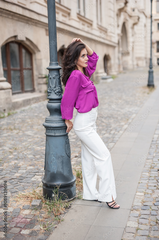 Beautiful young brunette woman in a purple blouse and white pants posing outdoor leans against a pillar .Portrait of a beautiful young brunette woman in a purple jacket posing