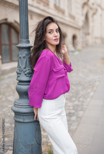 Obraz na płótnie Beautiful young brunette woman in a purple blouse and white pants posing outdoor leans against a pillar .Portrait of a beautiful young brunette woman in a purple jacket posing w salonie