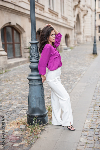 Obraz na płótnie Beautiful young brunette woman in a purple blouse and white pants posing outdoor leans against a pillar .Portrait of a beautiful young brunette woman in a purple jacket posing w salonie