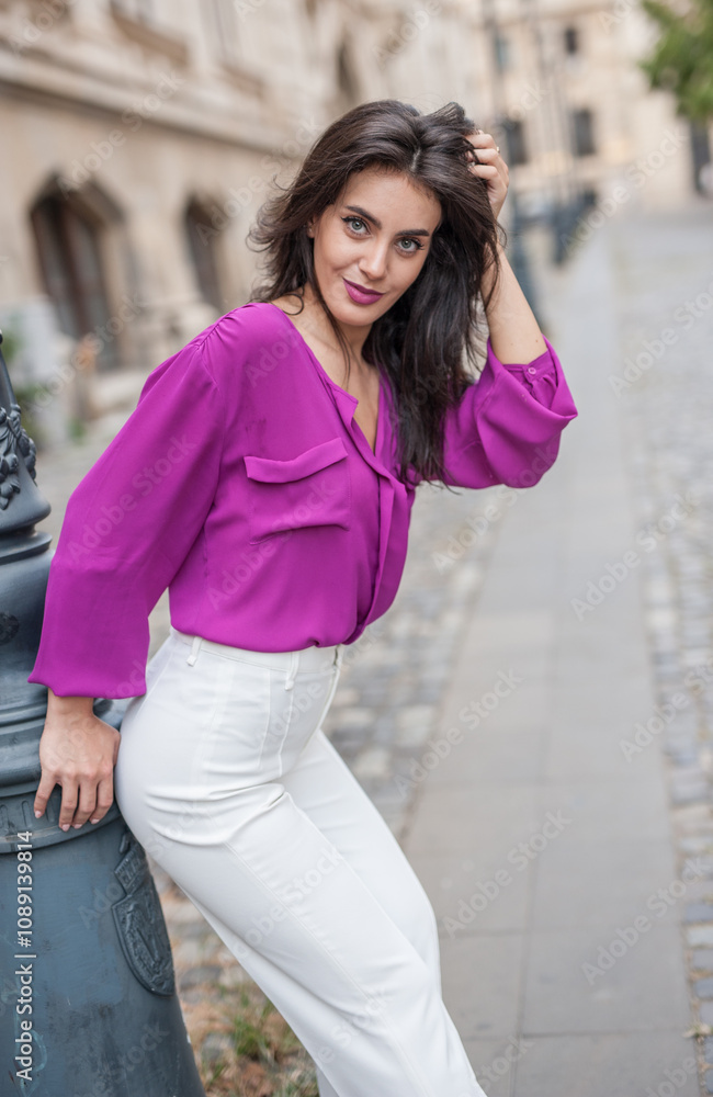 Beautiful young brunette woman in a purple blouse and white pants posing outdoor leans against a pillar .Portrait of a beautiful young brunette woman in a purple jacket posing