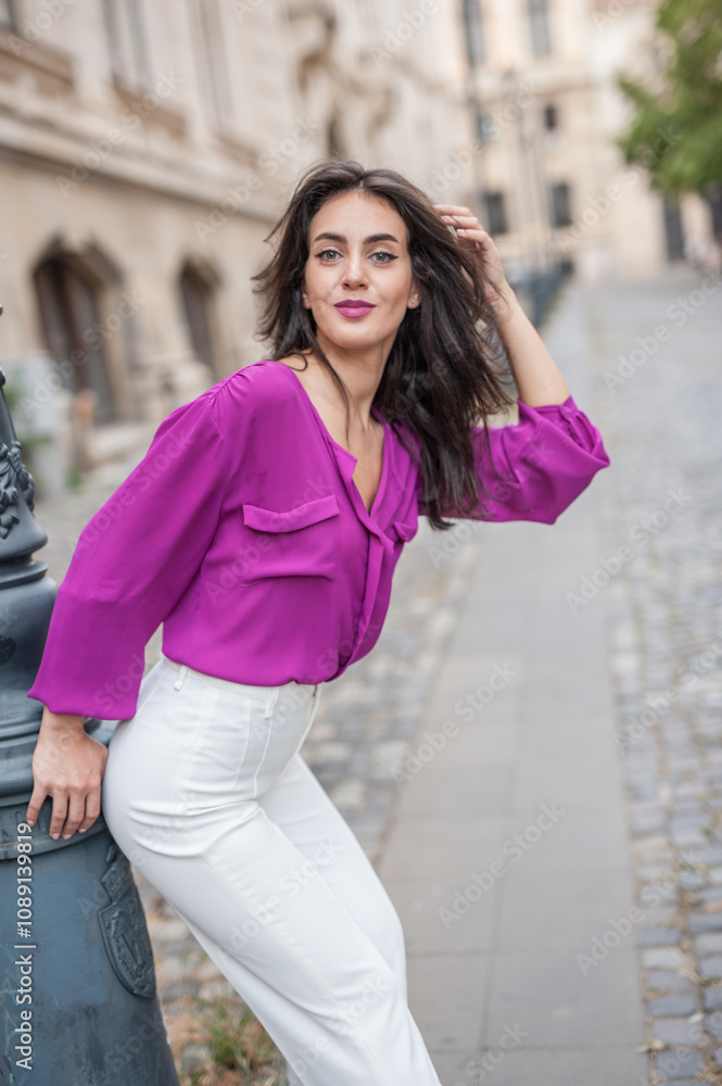 Beautiful young brunette woman in a purple blouse and white pants posing outdoor leans against a pillar .Portrait of a beautiful young brunette woman in a purple jacket posing