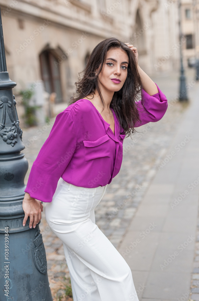 Beautiful young brunette woman in a purple blouse and white pants posing outdoor leans against a pillar .Portrait of a beautiful young brunette woman in a purple jacket posing