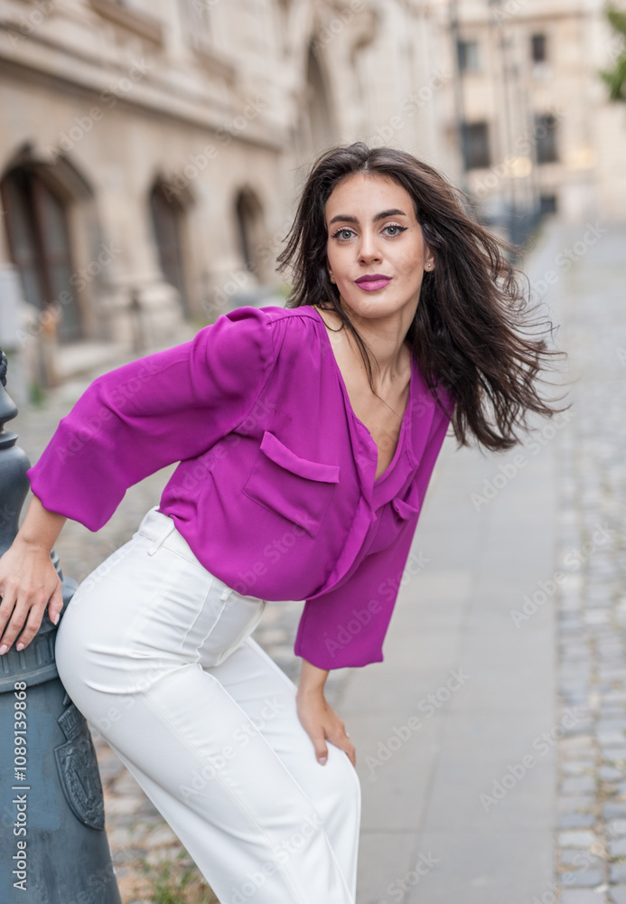 Beautiful young brunette woman in a purple blouse and white pants posing outdoor leans against a pillar .Portrait of a beautiful young brunette woman in a purple jacket posing
