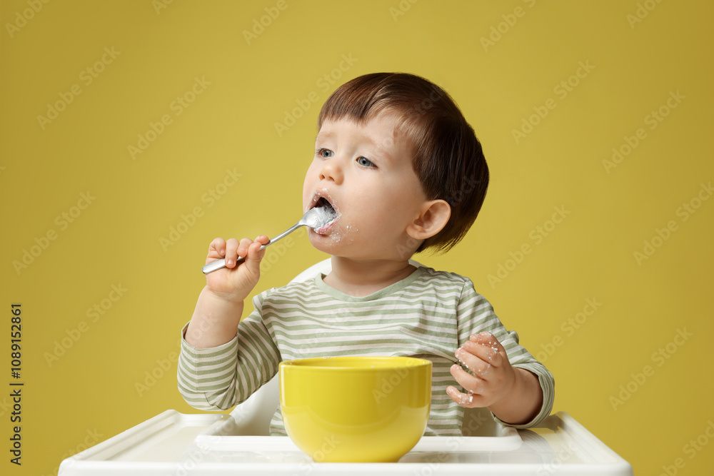 Cute little kid eating healthy baby food from bowl in high chair on yellow background Cute little kid eating healthy baby food from bowl in high chair on yellow background