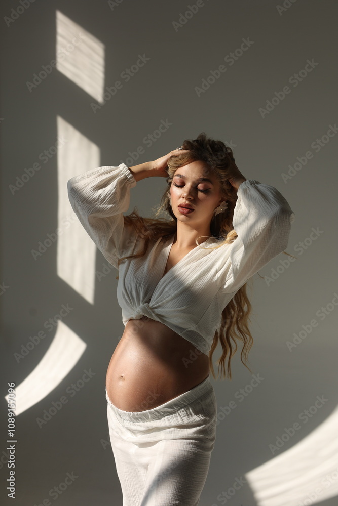 Fototapeta premium Beautiful pregnant woman in special white trousers and top. Advertisement of comfortable pregnancy Happy blond pregnant girl stand in studio with sun light. Long curly hair. Shadows on floor