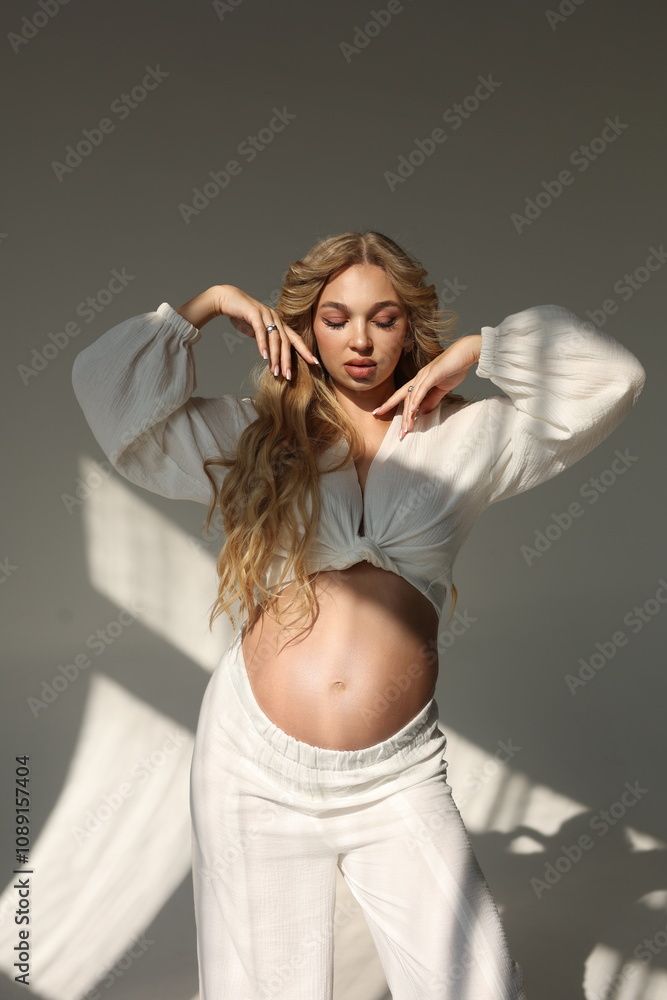 Fototapeta premium Beautiful pregnant woman in special white trousers and top. Advertisement of comfortable pregnancy Happy blond pregnant girl stand in studio with sun light. Long curly hair. Shadows on floor