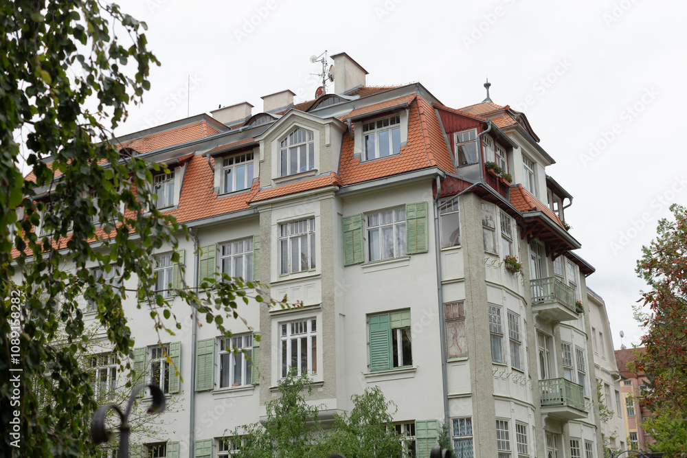 Fototapeta premium A charming European apartment building boasts a vibrant green facade adorned with decorative details and complemented by a striking red roof, all of which is beautifully surrounded by lush trees