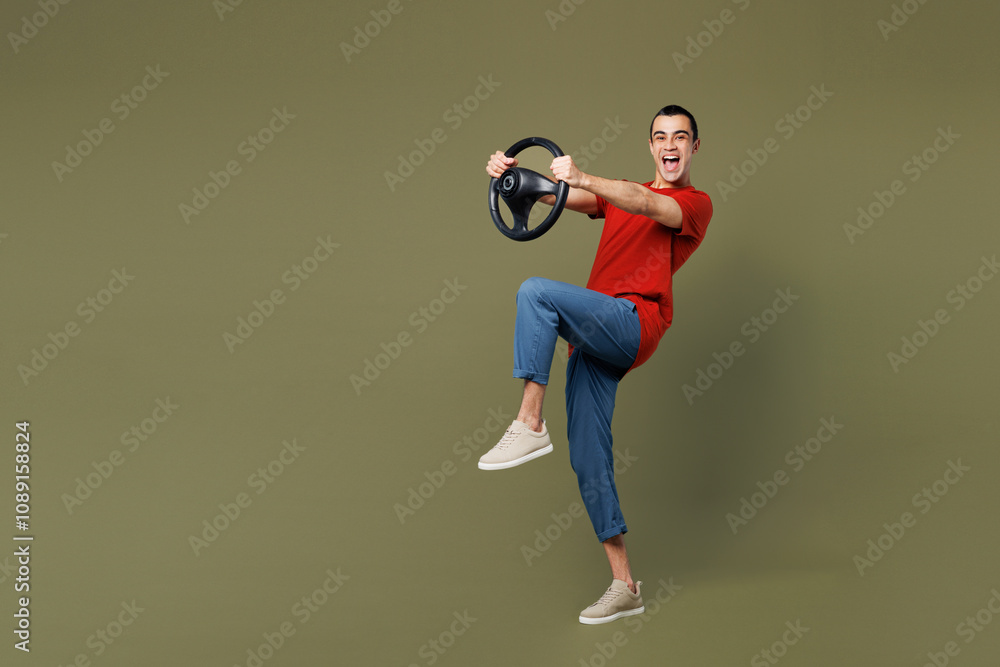Full body side view young middle eastern man wear red t-shirt casual clothes hold steering wheel driving car raise up leg isolated on plain pastel green background studio portrait. Lifestyle concept. Full body side view young middle eastern man wear red t-shirt casual clothes hold steering wheel driving car raise up leg isolated on plain pastel green background studio portrait. Lifestyle concept.