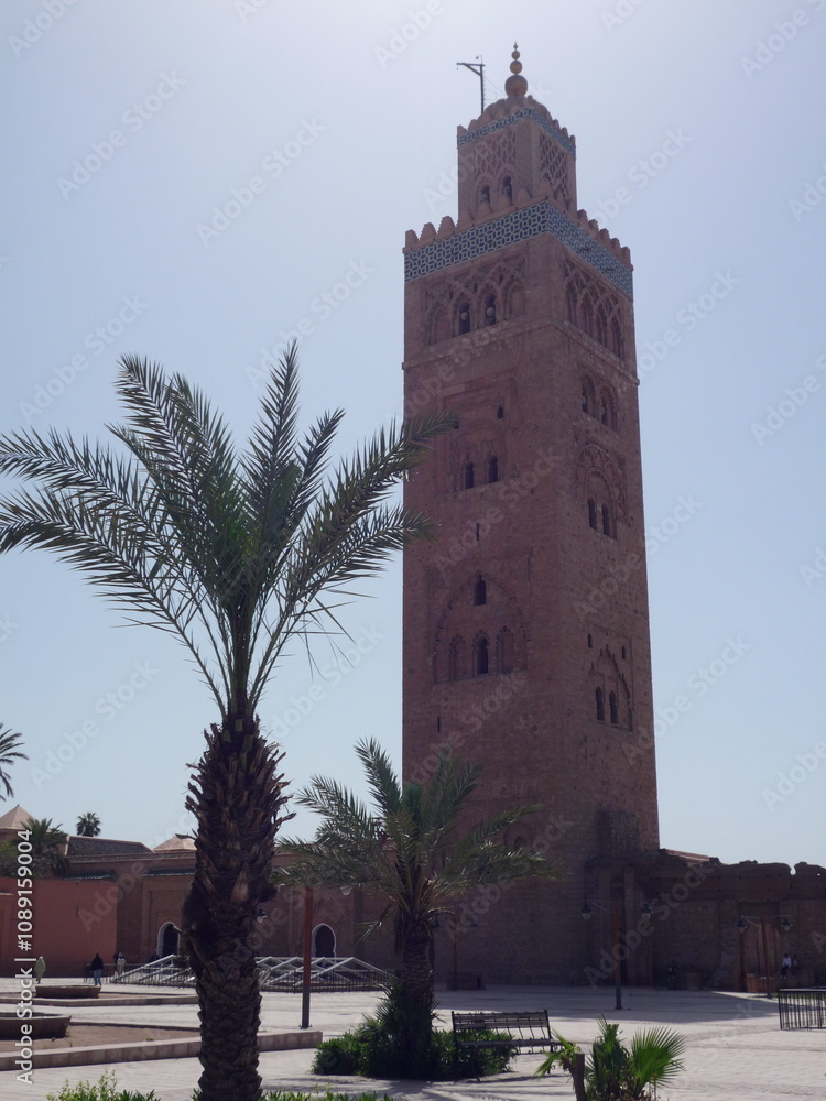 Fototapeta premium The iconic Koutoubia Mosque rises majestically against a bright blue sky, surrounded by swaying palm trees and a circular stone fountain in the heart of Marrakech.