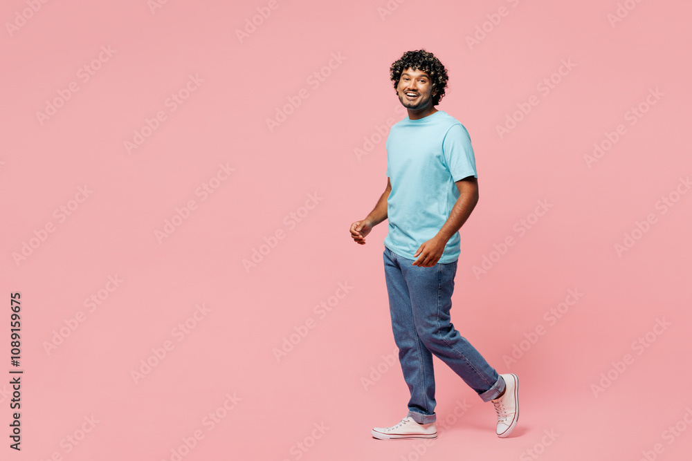 Full body side profile view young smiling Indian man he wear blue t-shirt casual clothes walking going looking camera isolated on plain pastel light pink background studio portrait. Lifestyle concept. Full body side profile view young smiling Indian man he wear blue t-shirt casual clothes walking going looking camera isolated on plain pastel light pink background studio portrait. Lifestyle concept.