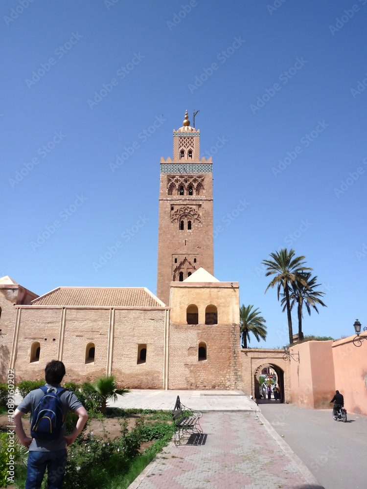 Obraz premium The Koutoubia Minaret, a Marrakech landmark, rises majestically above a lush garden, offering a striking contrast between the intricate red brickwork and the surrounding greenery against a clear blue 