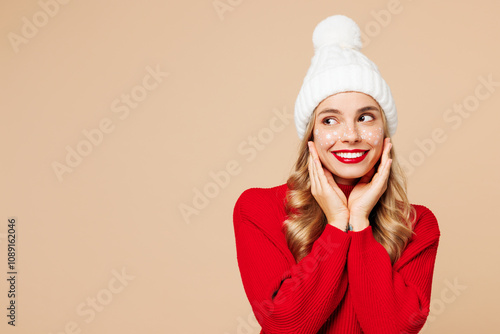 Merry cute young woman wears red warm cozy knitted sweater hat posing look aside put hands on face isolated on plain pastel light beige background Happy New Year celebration Christmas holiday concept photo