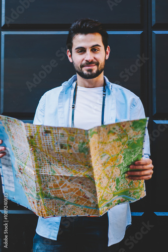 Half length portrait of handsome bearded tourist dressed in casual wear standing with opened travel map near copy space area on promotional background.Young man lover of trips smiling at camera photo