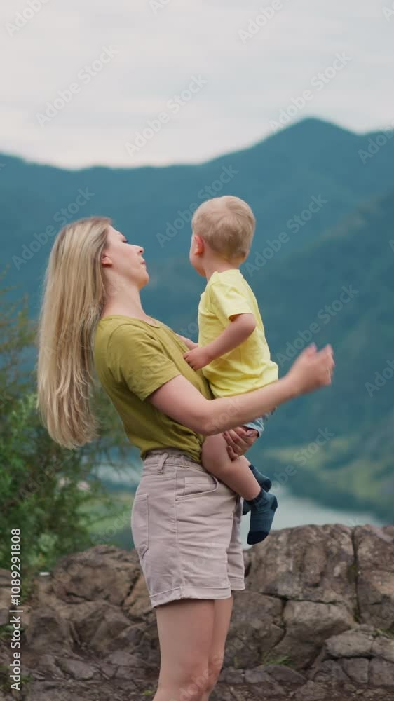 Young mother adjusts loose hair holding toddler boy in arms on rocky hill top against mountain silhouettes at highland on gloomy day slow motion Young mother adjusts loose hair holding toddler boy in arms on rocky hill top against mountain silhouettes at highland on gloomy day slow motion