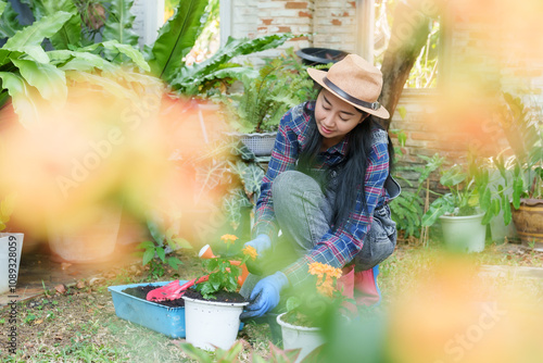 фотографія Asian woman in plaid shirt and overalls tending orange flowers in garden with soft focus foreground of vibrant foliage during sunny day showing tranquil and green environment