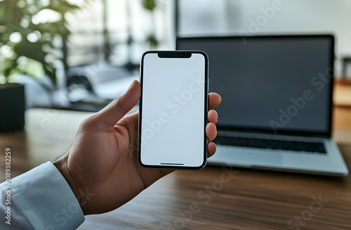 Close-up of a person's hand holding a smartphone with a blank screen, in front of a laptop on a wooden desk.